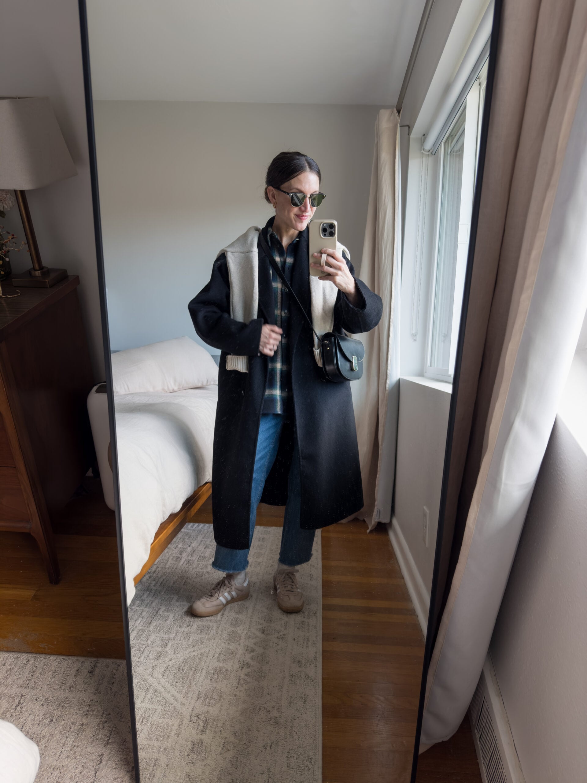 casual winter outfit with a plaid button down, straight leg jeans, tan Samba sneakers, black wool coat, beige sweater worn over the shoulders, and a black leather crossbody bag
