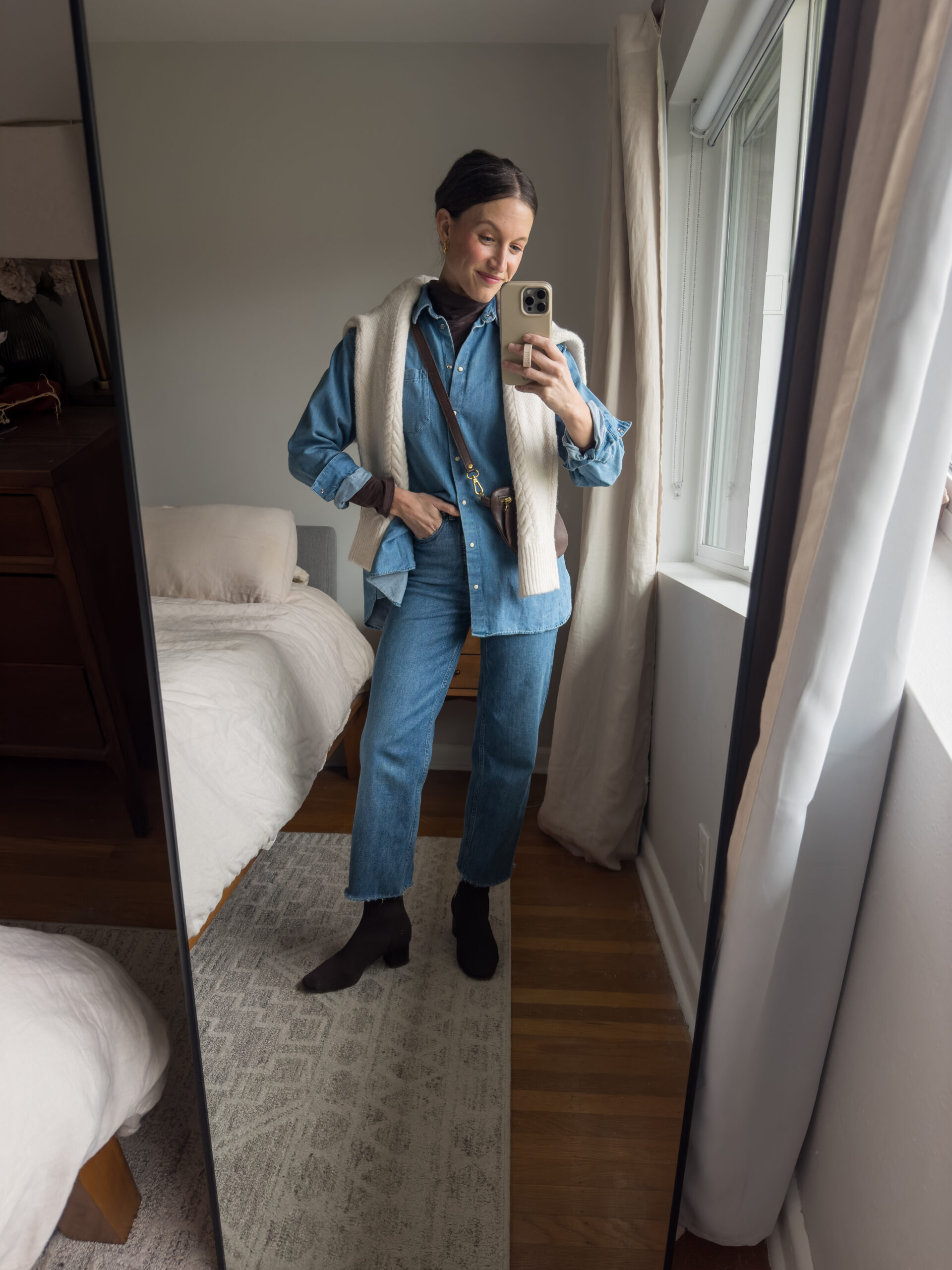 casual chic outfit idea with an oversized blue denim button down, a brown turtleneck, straight leg jeans, brown heeled boots, a cable knit sweater worn over the shoulders, and a brown sling bag