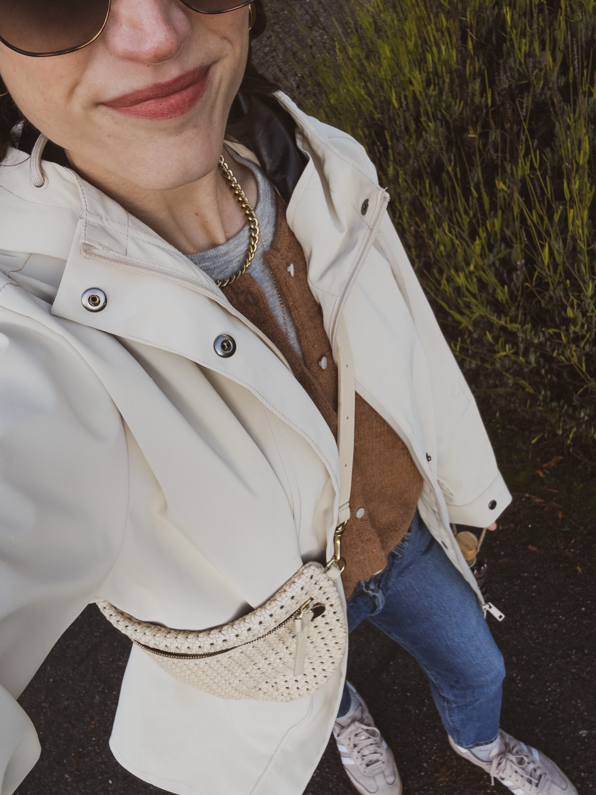 casual winter outfit with a gray tee, brown wool cardigan, beige rubber raincoat, straight leg jeans, tan Samba sneakers, and a cream sling bag