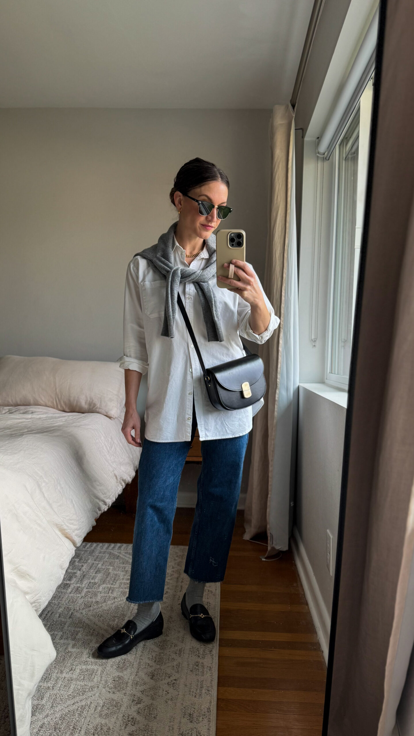 casual chic winter outfit with a white button down, gray sweater worn over the shoulders, dark wash straight leg jeans, black loafers with gray socks, and a black leather crossbody bag