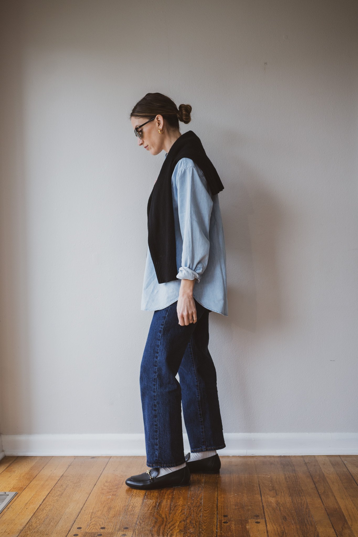 how to style loafers with a light denim button down and black sweater over the shoulders, dark wash jeans, off white socks, and black loafers
