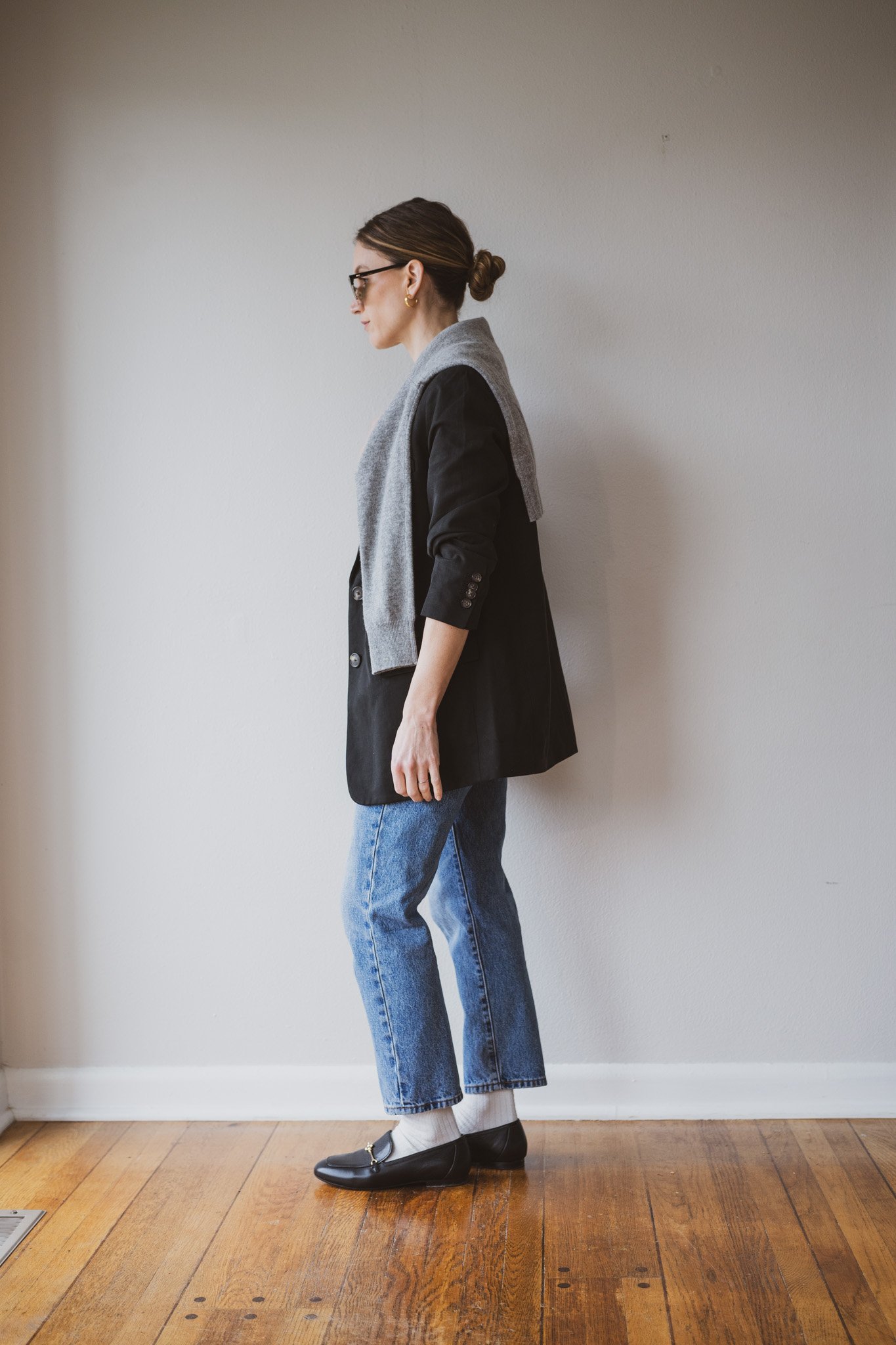 how to style loafers with a black blazer, gray sweater over the shoulders, slim cut light wash denim, off-white socks, and black loafers