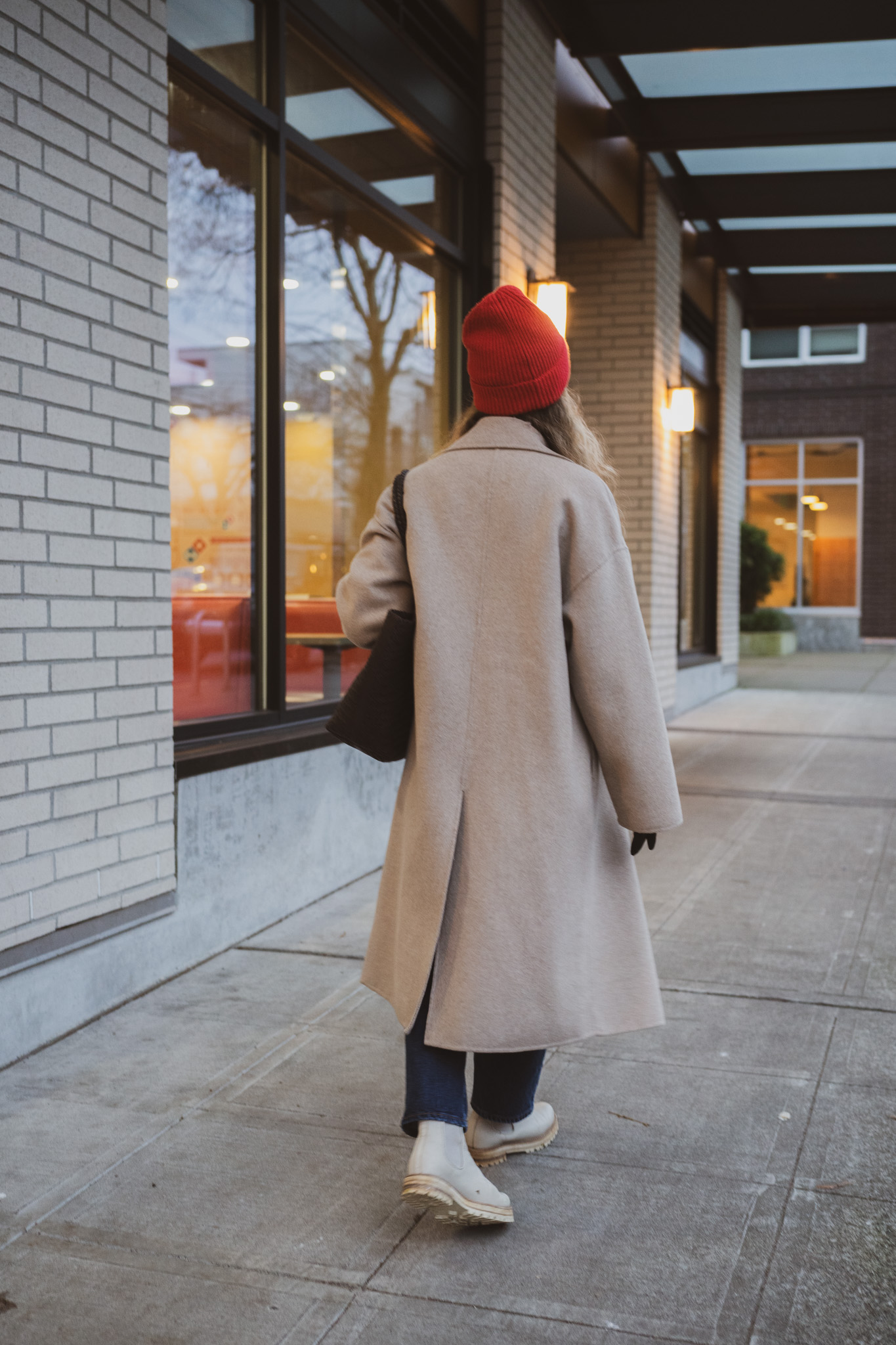 winter staples for cold weather, a cream colored cable knit sweater, tan wool coat, dark wash jeans, cream colored boots, and a red beanie