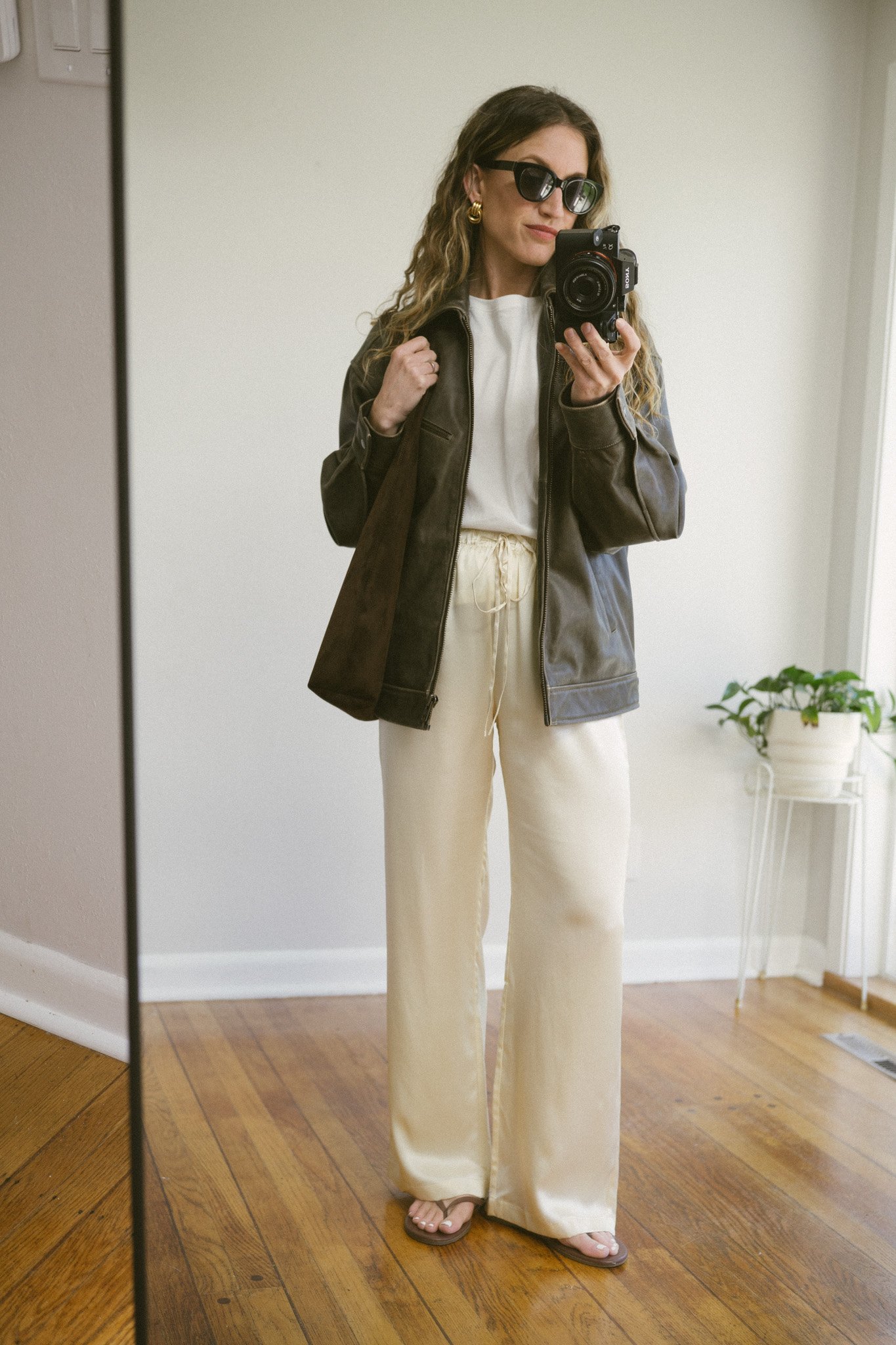 silk pants outfit idea with a white tee, brown leather bomber jacket, cream silk pants, and brown flip flops