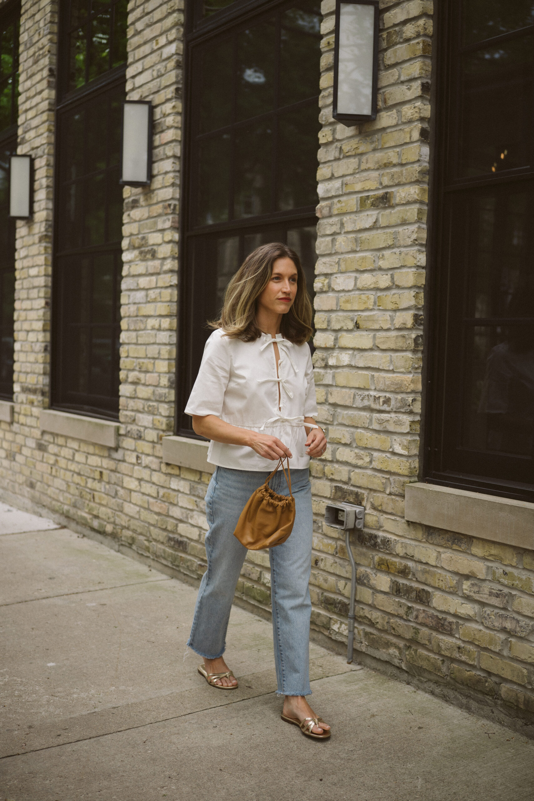 Quince rigid denim review - ashley wearing a white blouse with ties in the front, Quince rigid denim in a light wash, and gold sandals